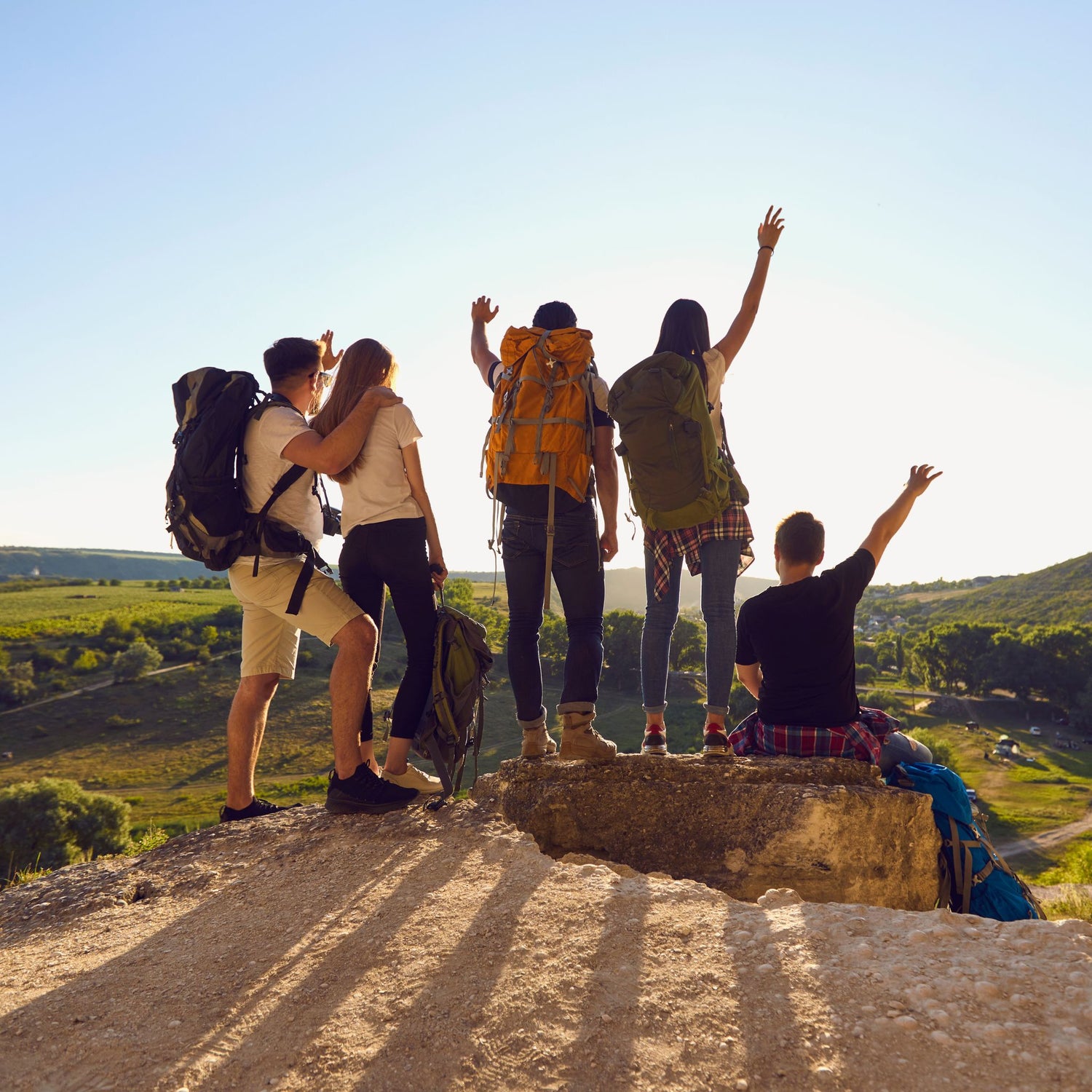 Group of hikers with backpacks standing on a rock with arms raised, overlooking a scenic landscape.