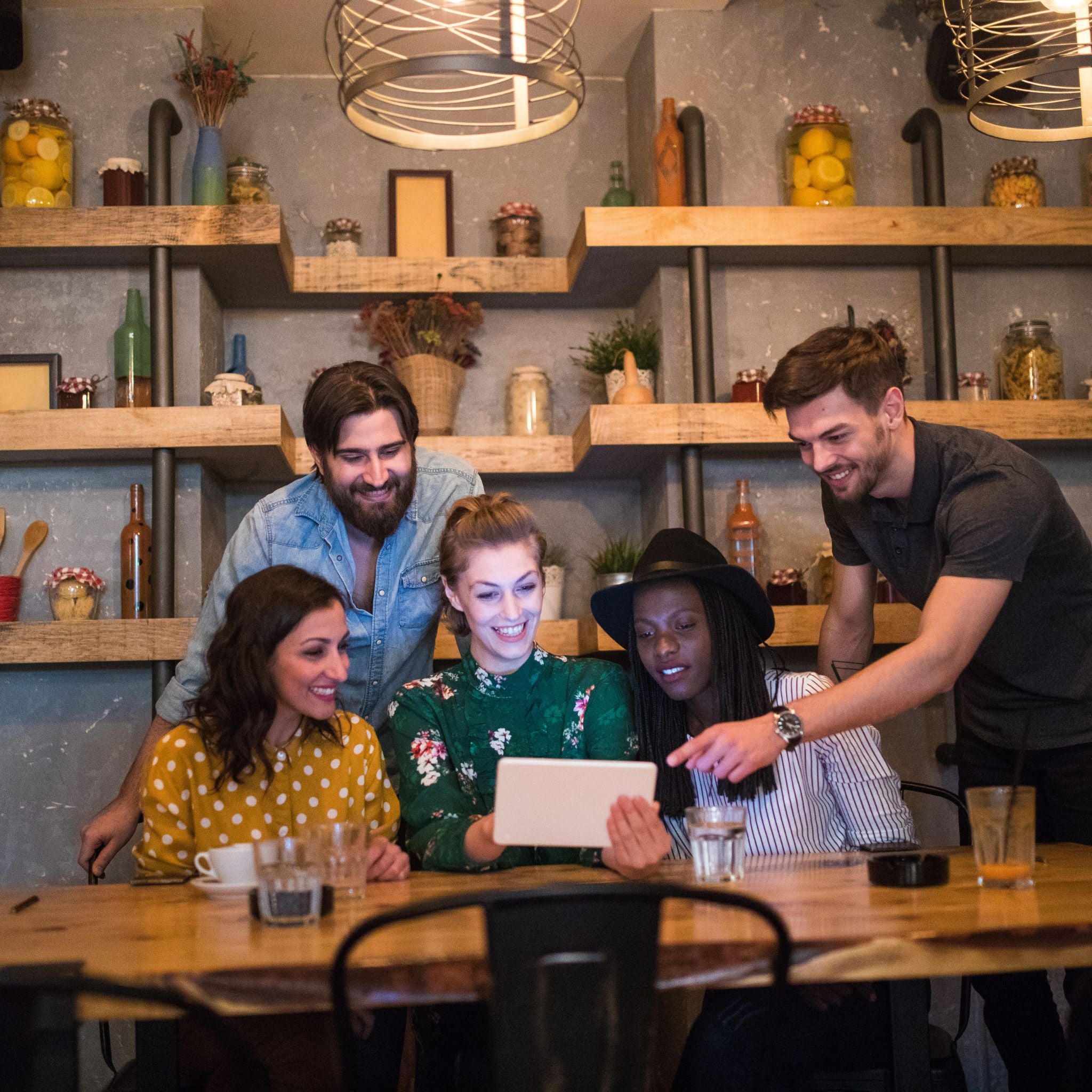 Group of friends gathered around a table in a restaurant, looking at a tablet and smiling as they look at an online memorial page.