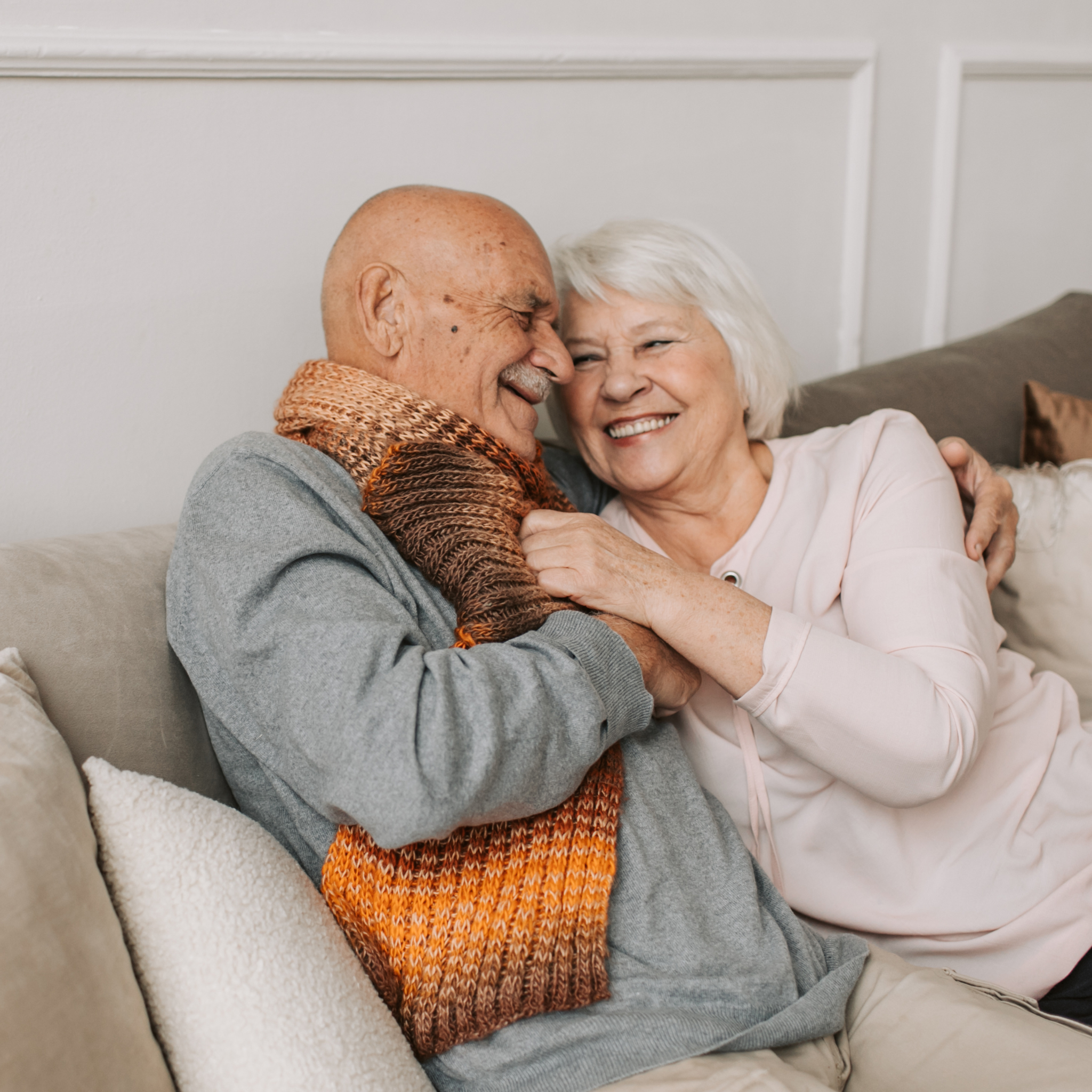Senior couple hugging on a couch, smiling. Creating memorial pages gently and with a lot of care.