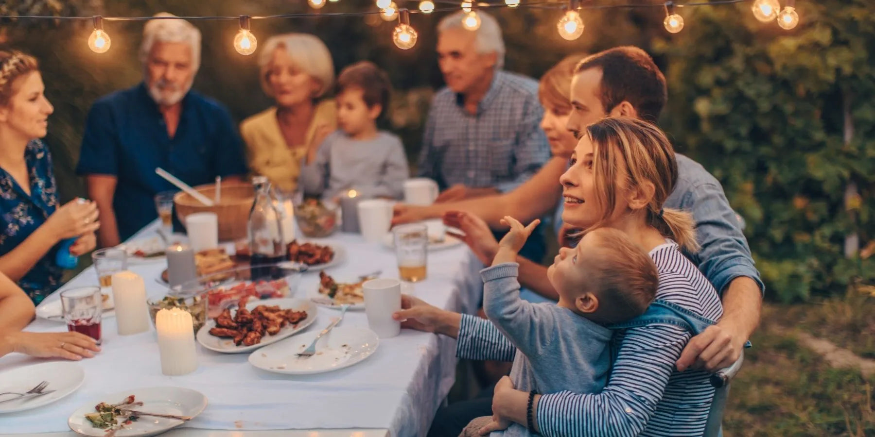 Family gathering around a dinner table outdoors at night with string lights.