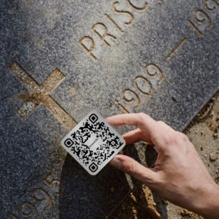 Hand holding a QR code card over a gravestone with engraved text.