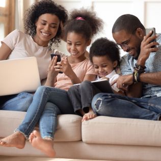 Family of four sitting on a couch, using electronic devices together.